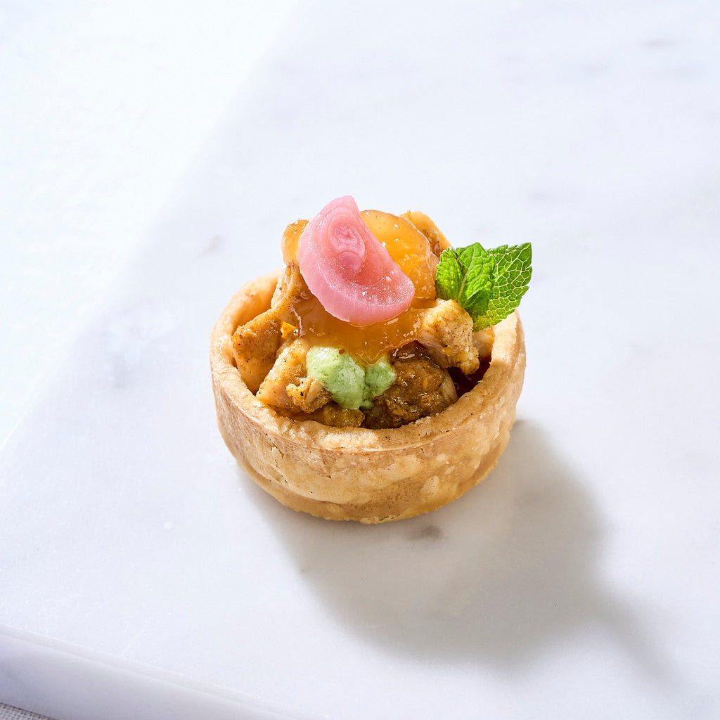 Single Chicken Tikka Tart with a pickled onion rose, mint leaf and green chutney on white marble