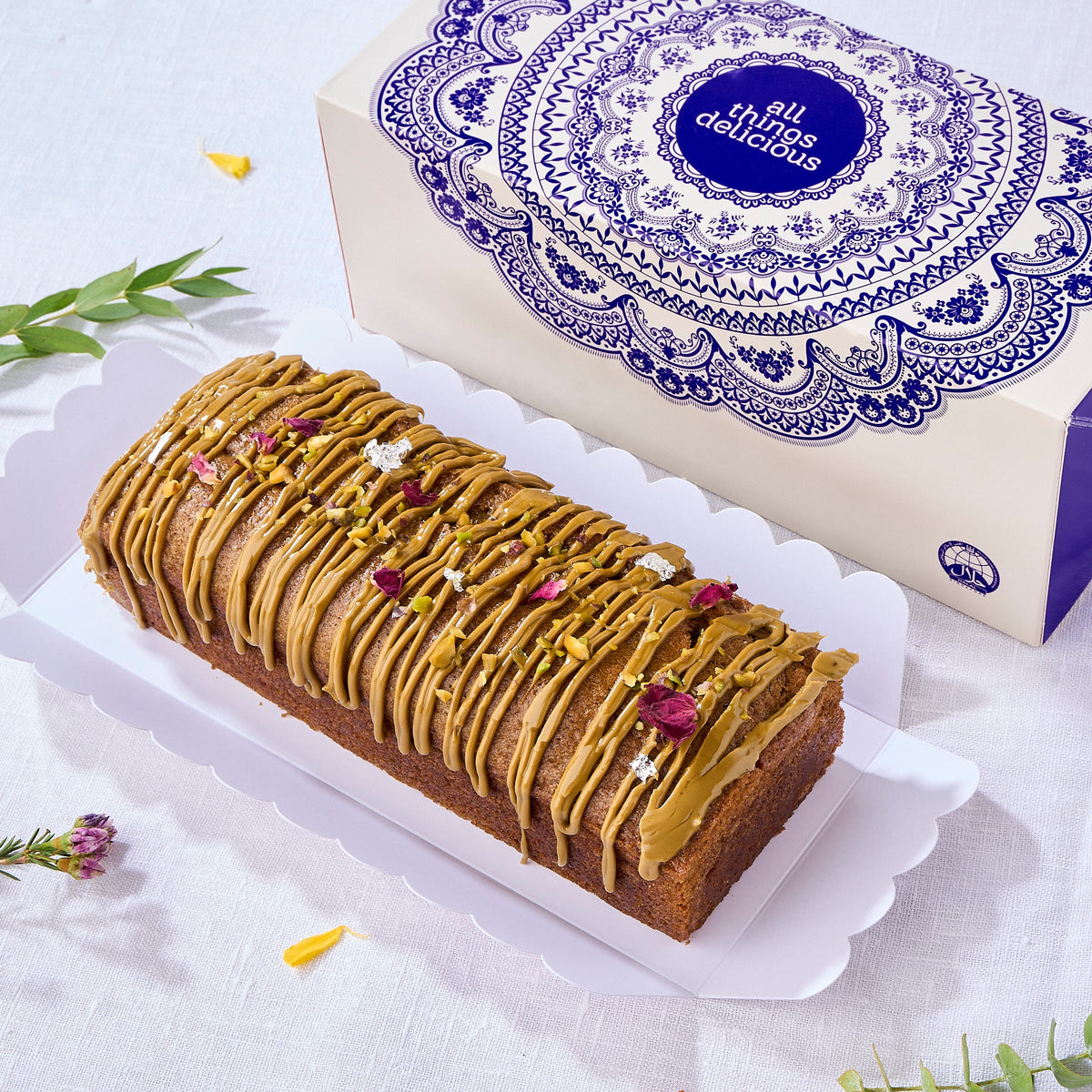 Eggless Pistachio Cardamom Voyage Cake topped with pistachio praline, rose petals and silver leaf by its gift box