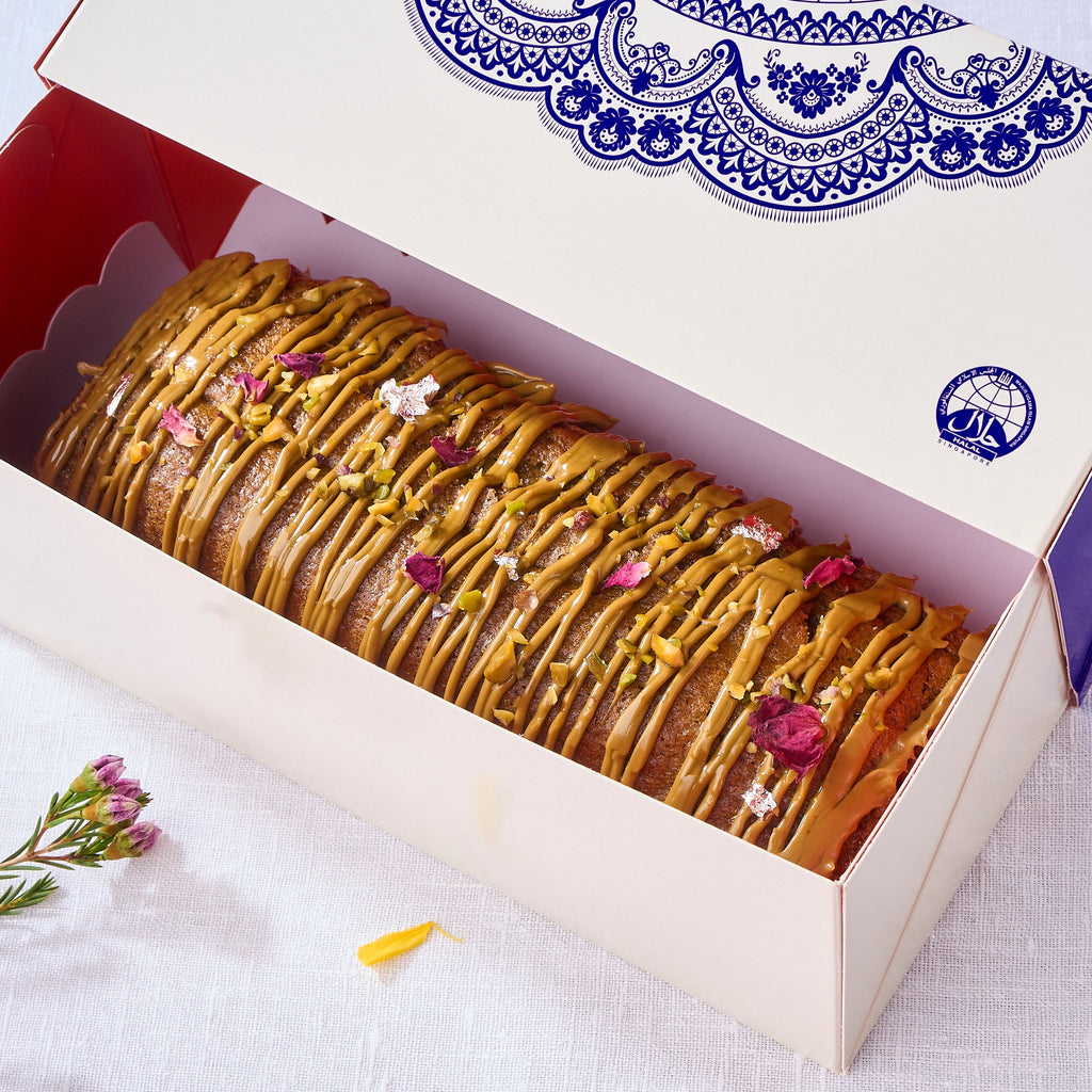 Pistachio Cardamom Voyage Cake in an open blue-patterned gift box showing a halal logo and praline drizzle